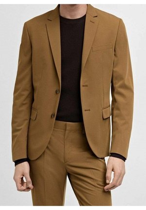 Mężczyzna ubrany w dopasowany brązowy garnitur z czarnym prążkowanym swetrem z okrągłym dekoltem pod spodem, stojący na tle jednolitego tła.