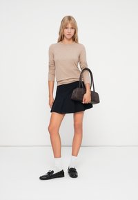 Beżowy sweter dzianinowy, czarna spódnica z plisami, białe skarpetki i czarne mokasyny. Brązowa torebka trzymana w jednej ręce, gładka faktura, minimalistyczny design.