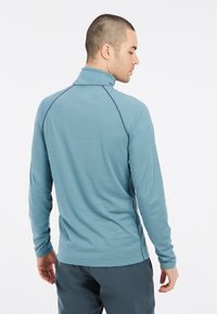 Maglietta attiva a maniche lunghe di colore turchese, con colletto alto, tessuto testurizzato e cuciture a contrasto. Presenta un design aderente con maniche raglan.