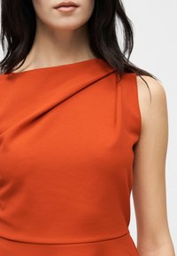 Γυναίκα που φοράει αμάνικο φόρεμα σε χρώμα καμένο πορτοκαλί με ασύμμετρο ντεκολτέ και απαλά πτυχώσεις.