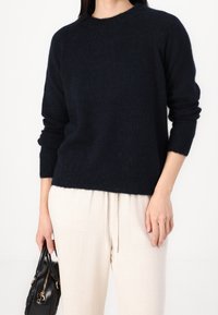Pull en maille bleu marine avec un col rond et des manches longues, porté avec un pantalon en lin beige clair. La personne tient un petit sac à main noir.