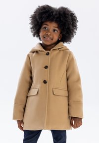 Manteau en polaire beige avec une capuche, comportant quatre boutons noirs, deux poches avant et une coupe droite. Le tissu semble doux et texturé.