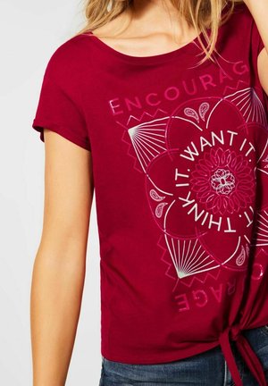 Rotes Baumwoll-T-Shirt mit kurzen Ärmeln, Rundhalsausschnitt und geknotetem Saum. Grafik auf der Vorderseite zeigt ein Blumenmotiv mit dem Text "DENKE ES, WILL ES, ERMUTIGE."