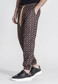 Pantalones de chándal marrones con un diseño estampado, una cinturilla con cordón y bolsillos laterales, que presentan un diseño de logo repetido. Combinados con zapatillas negras.
