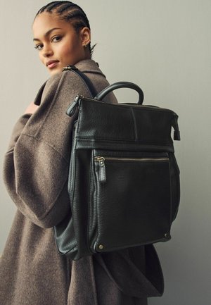 Schwarzer Leder-Rucksack mit strukturiertem Design. Verfügt über einen Handgriff, Reißverschlussfächer und Messingakzente. Wird über einem braunen Pullover getragen.