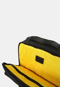 Borsa nera realizzata in materiale impermeabile, con un interno giallo brillante, scomparti con cerniera e un'etichetta in pelle con il marchio.