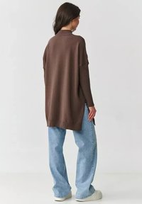 Brązowy, oversize sweter z wysokim kołnierzem, prążkowaną fakturą i bocznymi rozcięciami, w połączeniu z jasnoniebieskimi, luźnymi dżinsami i białymi butami.