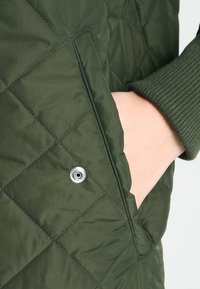 Dark green quilted jacket featuring a side pocket with a snap closure. The sleeve has ribbed knit detailing for added texture.