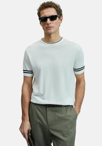 Man met zwarte zonnebril, witte shirt met korte mouwen en zwarte strepen op de mouwen, en olijfgroene broek, die een bruin voorwerp vasthoudt.