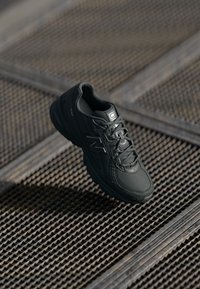 Fekete sportcipő, texturált szintetikus felsőrésszel, párnázott talppal és fűzőkkel, rácsos fém felületen elhelyezve.