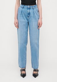 Μπλε τζιν παντελόνι ψηλής μέσης με πιέτες, ίσια γραμμή και ορατά ραψίματα. Συνδυασμένο με μαύρες γόβες με καμπυλωτή μύτη και λουράκια.