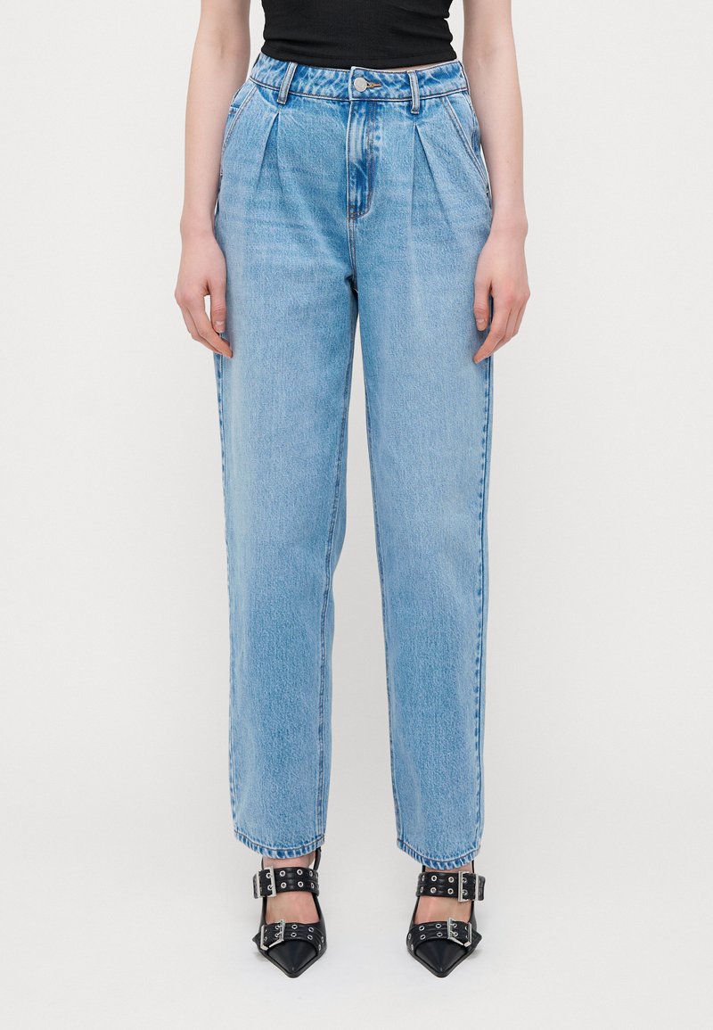 Μπλε τζιν παντελόνι ψηλής μέσης με πιέτες, ίσια γραμμή και ορατά ραψίματα. Συνδυασμένο με μαύρες γόβες με καμπυλωτή μύτη και λουράκια.