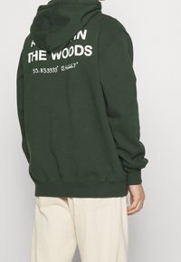 Felpa verde scuro con una grande grafica bianca sul retro che recita "DA SOLO NELLE FORESTE" e le coordinate. Tessuto morbido e strutturato, vestibilità relaxed.