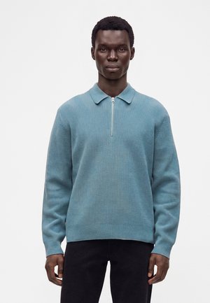 Man met een donkere huid die een blauw geribbelde trui met rits en zwarte broek draagt, staand voor een effen witte achtergrond.