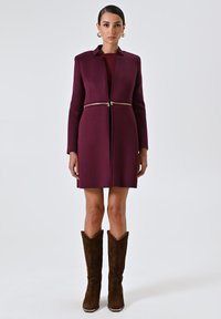 Cappotto tailored bordeaux con colletto e dettagli in zip dorata, indossato sopra un top coordinato, abbinato a stivali da cowboy marroni ricamati.