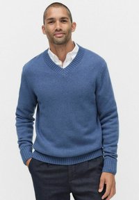 Niebieski sweter w serek z żeberkowymi mankietami i dołem, wykonany z miękkiego materiału. Noszony na białej koszuli z kołnierzykiem, stylizowany z ciemnymi spodniami.