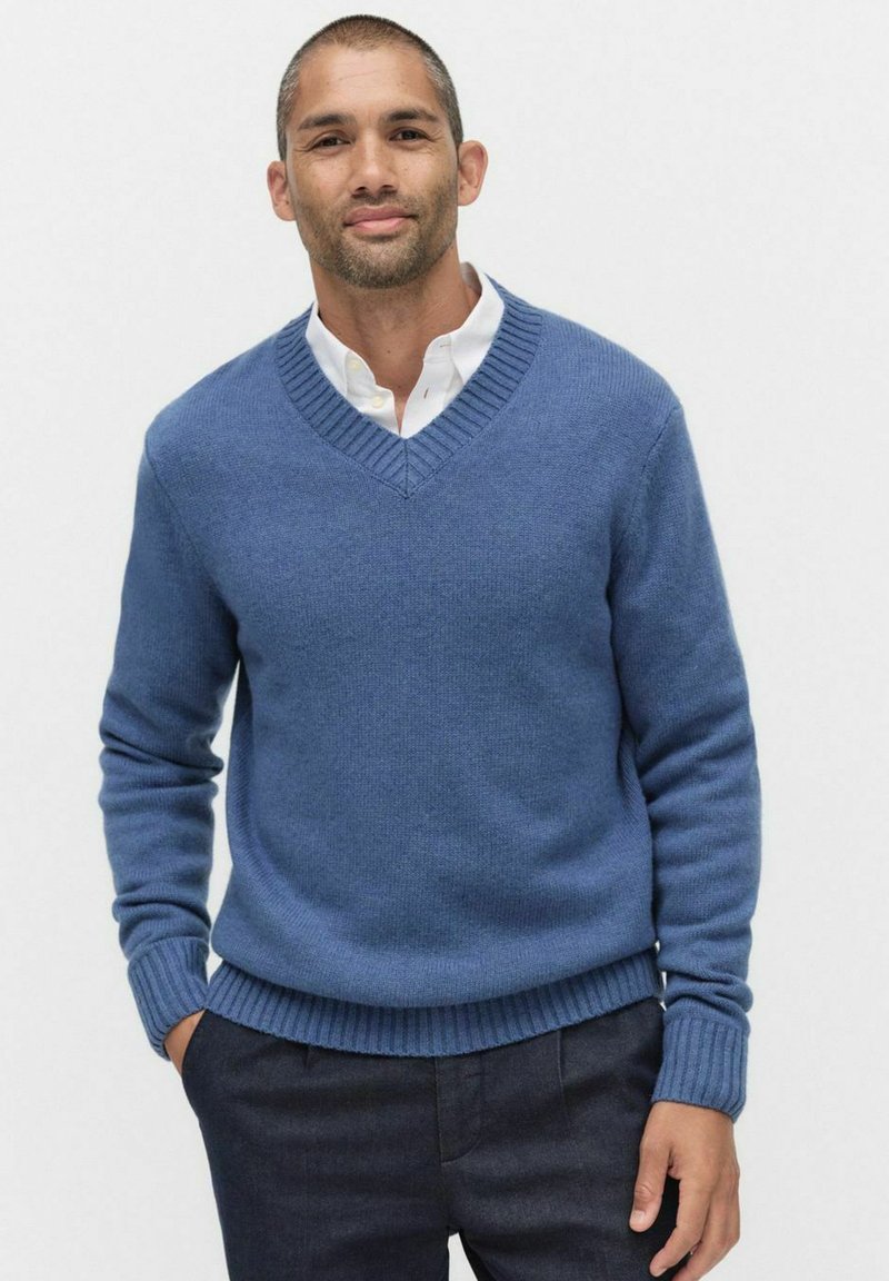 Niebieski sweter w serek z żeberkowymi mankietami i dołem, wykonany z miękkiego materiału. Noszony na białej koszuli z kołnierzykiem, stylizowany z ciemnymi spodniami.