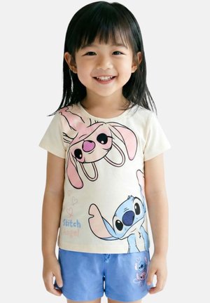 Jeune fille souriante, portant un t-shirt crème avec des personnages de dessins animés roses et bleus et un short bleu assorti avec un design similaire.
