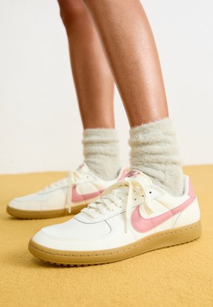 Ben iført hvide sneakers med pink Nike-swoosh og beige såler, kombineret med lysegrå, lodne sokker på gul gulv.