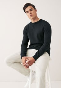 Suéter negro de punto con mangas largas y cuello redondo, acompañado de pantalones beige claros y zapatillas blancas, sentado en un taburete blanco.