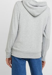 Persona con una sudadera con capucha gris claro y jeans de mezclilla azul, vista desde atrás, de pie contra un fondo claro y liso.
