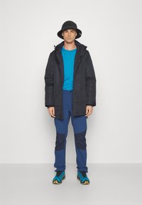 Svart isolerad jacka, blå kortärmad skjorta, tvåtonade blå byxor med knälappar och flerfärgade sportskor. Svart bucket hat.