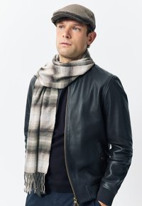 Leather bomber jacket in black, featuring a zip front and side pockets. Wrapped in a light plaid scarf with fringe. Gray flat cap worn.