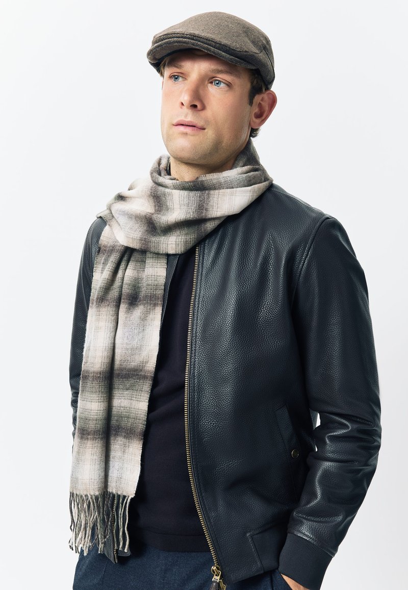 Leather bomber jacket in black, featuring a zip front and side pockets. Wrapped in a light plaid scarf with fringe. Gray flat cap worn.