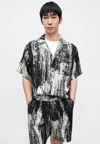 Komplet s kratkimi rokavi, ki vključuje črno-belo srajco z vzorcem in ujemajoče se kratke hlače, narejen iz lahkotne tkanine, z gumbi na sprednji strani in dvema žepoma.