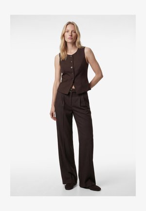 Kobieta stojąca z ręką za plecami, ubrana w bezrękawnik w ciemnobrązowym kolorze z guzikami oraz pasujące szerokie spodnie na tle białego tła.