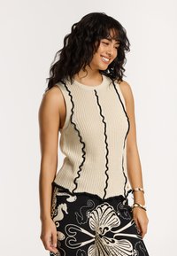 Adīts bez piedurknēm tops krēmkrāsā ar melnām ripojošām apakšmalām un pieguļošu formu. Valkāts ar rakstainām melnbaltām biksēm.