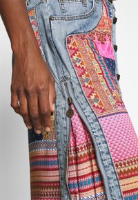 Mörkhyad hand som vilar på höften nära en flerfärgad lapptäcksjeanskjol med en gyllene dragkedja och intrikata mönster.
