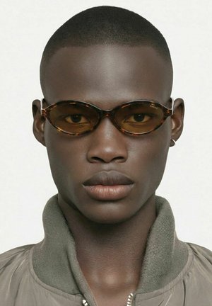 Jeune homme noir aux cheveux très courts portant des lunettes de soleil ovales écaille de tortue et une veste bomber verte, sur un fond blanc uni.