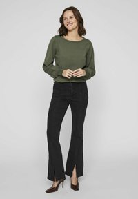 Zaļa adīta džemperis ar apaļu kakla izgriezumu un pūkainām piedurknēm, kopā ar melnām platām biksēm ar šķeltiem apakšējiem galiem un brūnām norādītas augstpapēžu kurpēm.