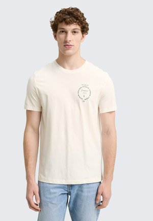 Cremefarvet bomuld t-shirt med rund halsudskæring og korte ærmer. Har et trykt logo på venstre brystområde. Bæres sammen med lyseblå jeans.