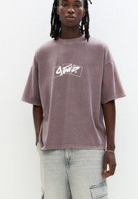 T-shirt oversize di colore malva in morbido cotone, con una grafica bianca sul petto, maniche corte e una vestibilità comoda.