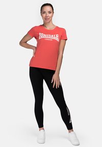 Red short-sleeve t-shirt with a white graphic "Lonsdale London" print, paired with black leggings featuring a white logo accent, and white sneakers.