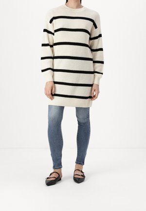 Persoon die een lange crèmekleurige trui met zwarte horizontale strepen draagt, blauwe strakke jeans en zwarte platte puntige schoenen met enkelbandjes.