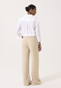 Camisa blanca de botones con pliegue en la espalda, combinada con pantalones de pierna ancha beige y bolsillos traseros. Tela suave con una textura sutil.
