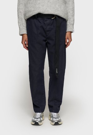Pantaloni in cotone blu navy con vestibilità rilassata, dotati di due tasche frontali e una cintura nera con etichetta brandizzata. Sneakers argentate visibili in basso.