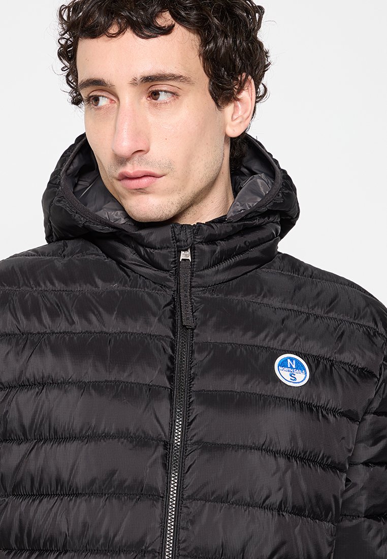 Giacca a vento nera con cappuccio, dotata di un motivo trapuntato, con zip frontale e una toppa blu sul lato sinistro del petto.