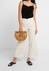 Kvinna i svart linne och krämfärgade vida byxor, som håller en halvcirkelformad handväska i bambu, och har svarta sandaler med nitar.