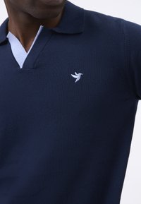 Námořnická modrá bavlněná polo košile s bílým límcem a malým vyšitým logem kolibříka na levé hrudi. Hladká textura, klasický střih.