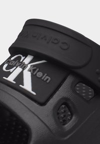 Μαύρο σανδάλι slide Calvin Klein με ρυθμιζόμενο λουράκι που διαθέτει ανάγλυφο λογότυπο Calvin Klein και οπές αερισμού.