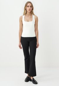 Vrouw staat in een witte mouwloze top met vierkante halslijn, zwarte flare-broeken en zwarte instapschoenen tegen een neutrale witte achtergrond.