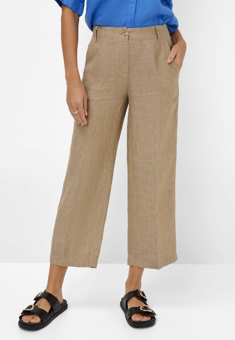 Lichtbruine linnen broek met wijde pijpen en een gladde textuur, voorzien van een knoopsluiting en zijzakken, gecombineerd met zwarte sandalen en een blauwe shirt.
