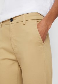 Gros plan d'une personne portant un pantalon beige avec la main dans la poche avant et une chemise blanche rentrée.