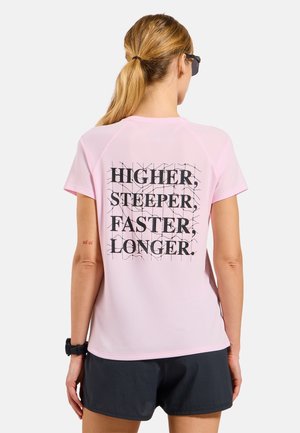 Frau mit blondem Pferdeschwanz, die eine Sonnenbrille, ein pinkes T-Shirt mit dem Schriftzug „Higher, Steeper, Faster, Longer“ auf dem Rücken und schwarze Shorts trägt.