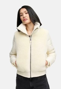 Pufferjack in crèmekleur met een fuzzy textuur aan de voorkant, geribbelde manchetten, hoge kraag en een zilveren rits. Draagt een zwarte top en donkere jeans.