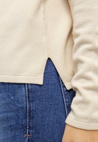 Beige Strickpullover mit abgerundetem Saum und seitlichen Schlitzen, kombiniert mit blauen Jeans. Weiche Textur und sichtbare saubere Nähte.